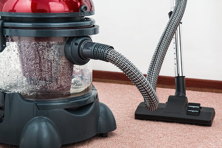 A woman using a yellow vacuum cleaner with a long hose on a patterned area rug with floral motifs and a beige background, situated on a wooden floor in a living room. She is dressed in casual clothing