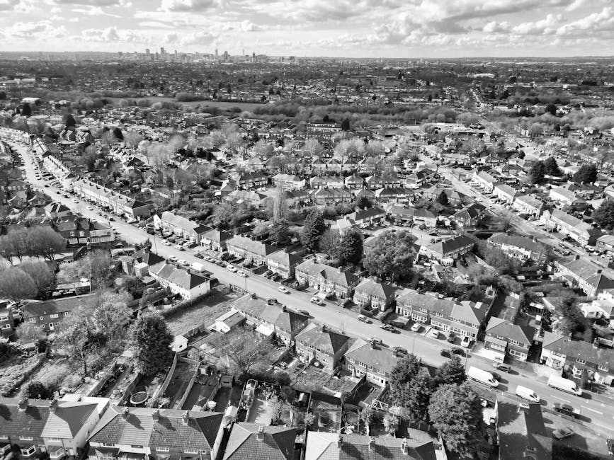 Black and white aerial photograph of a residential neighborhood showing rows of houses along tree-lined streets. The houses have pitched roofs, some with chimneys, and small gardens in front. The stre